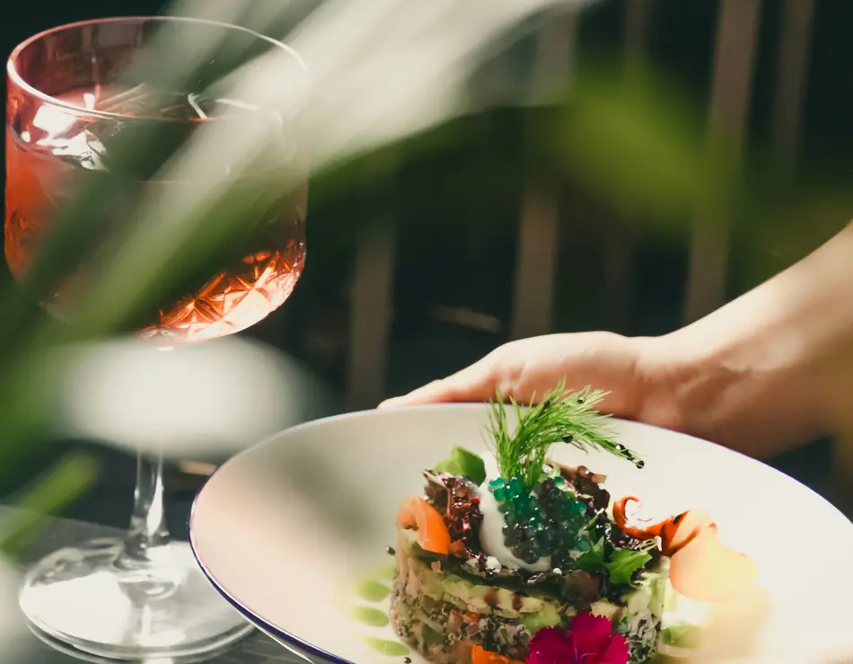 glockna, dein Genusshotel im SalzburgerLand Hände halten Teller mit kunstvoll angerichtetem Gourmetgericht neben einem Glas Getränk