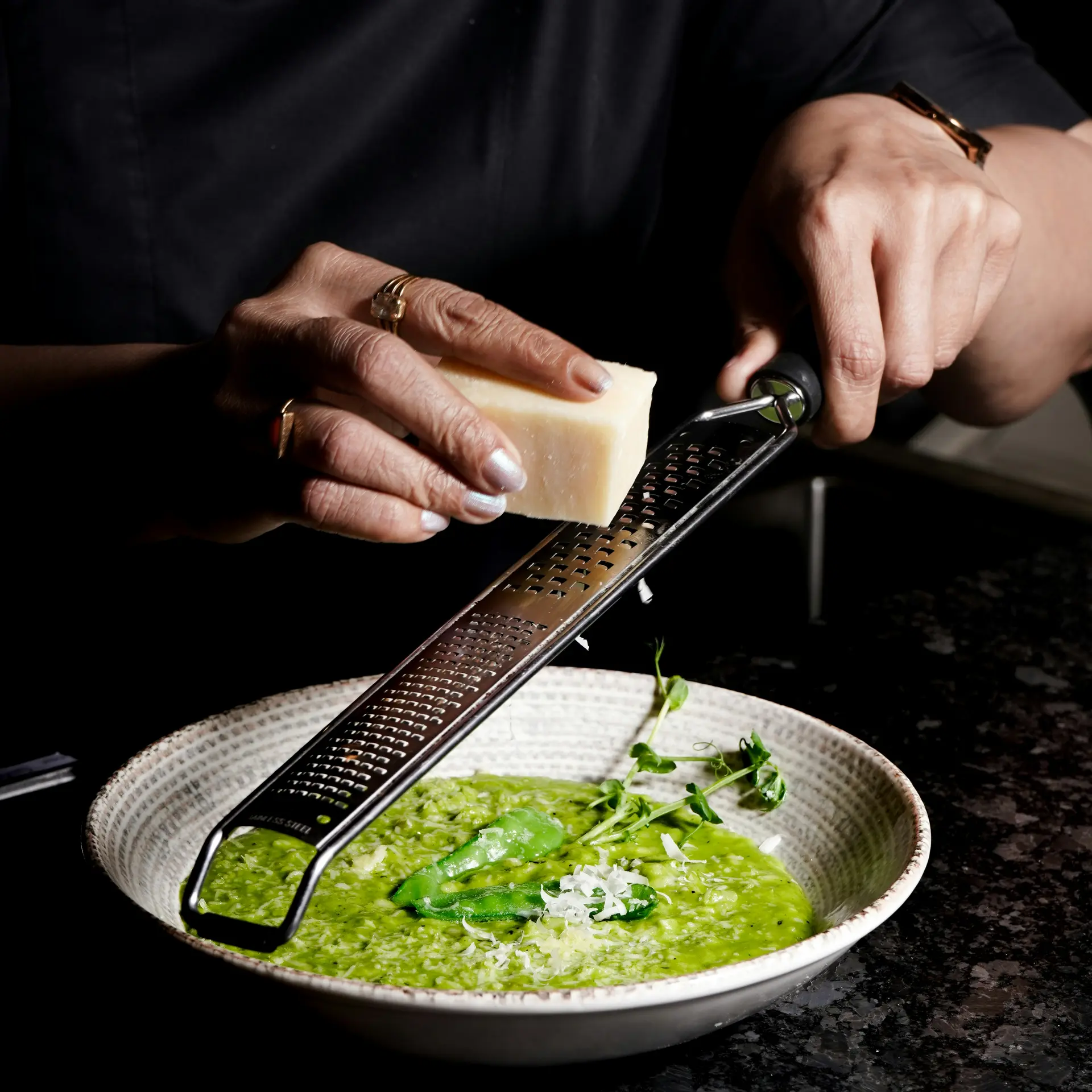 Person reibt Käse über grünes Risotto in weißem Teller