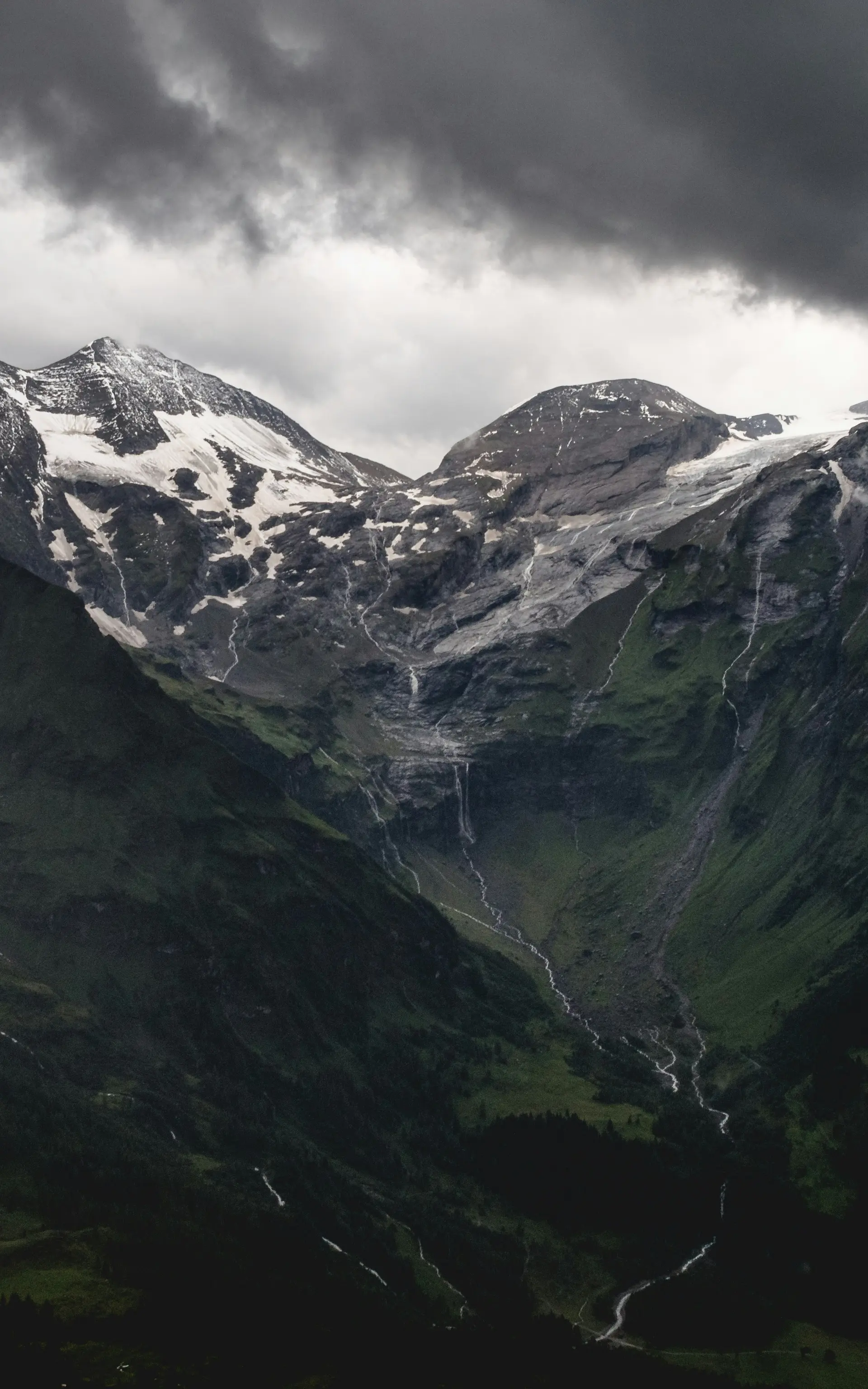Dunkle Wolken über schneebedeckten Berggipfeln und grünen Hängen
