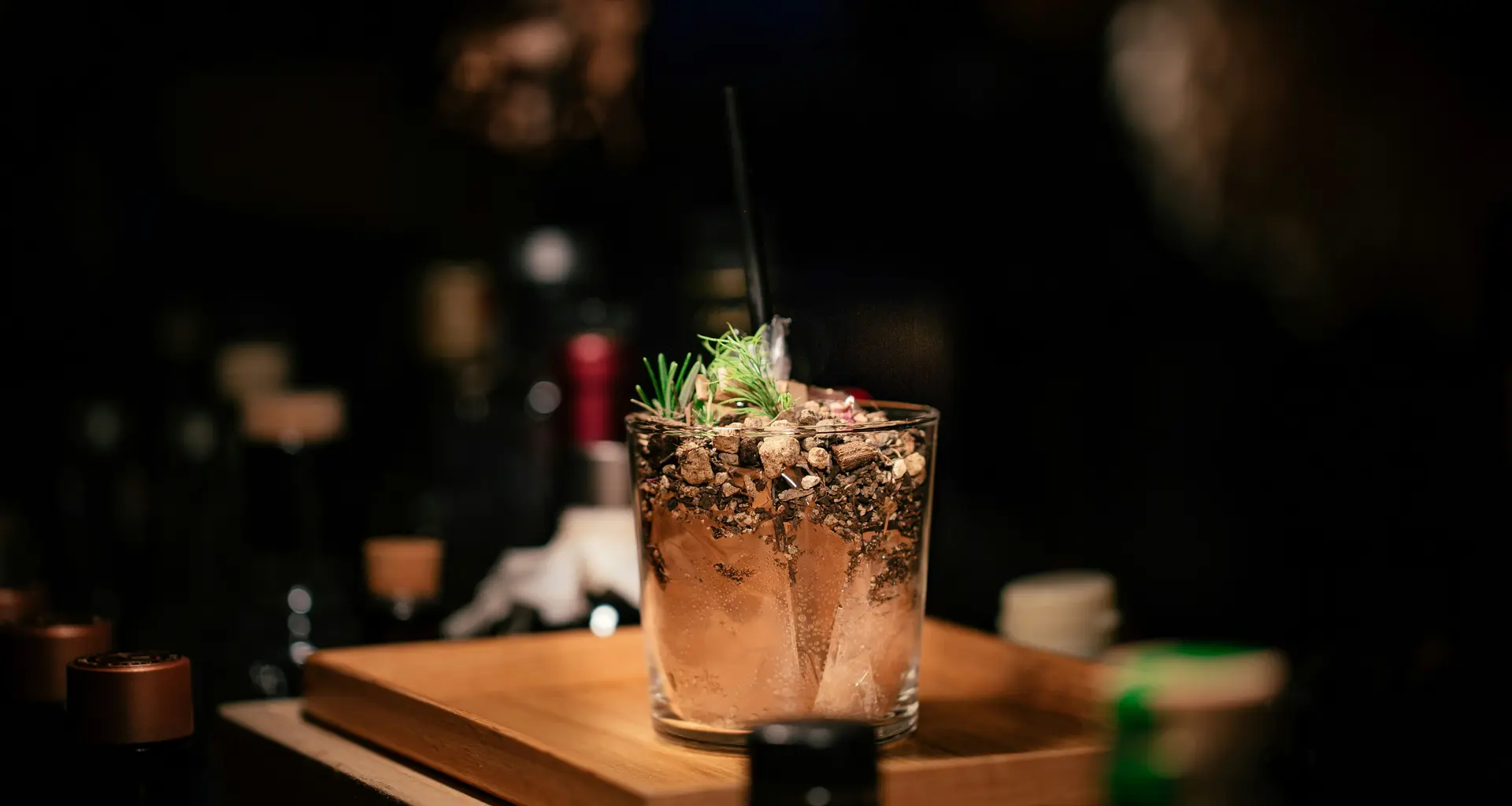 Cocktailglas mit Eis und Kräutern auf einem Holzbrett in dunkler Bar