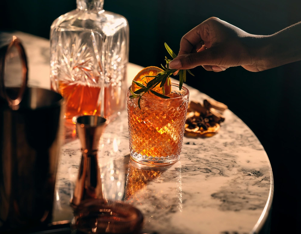 Hand garnishing cocktail with orange slice and herbs on marble surface