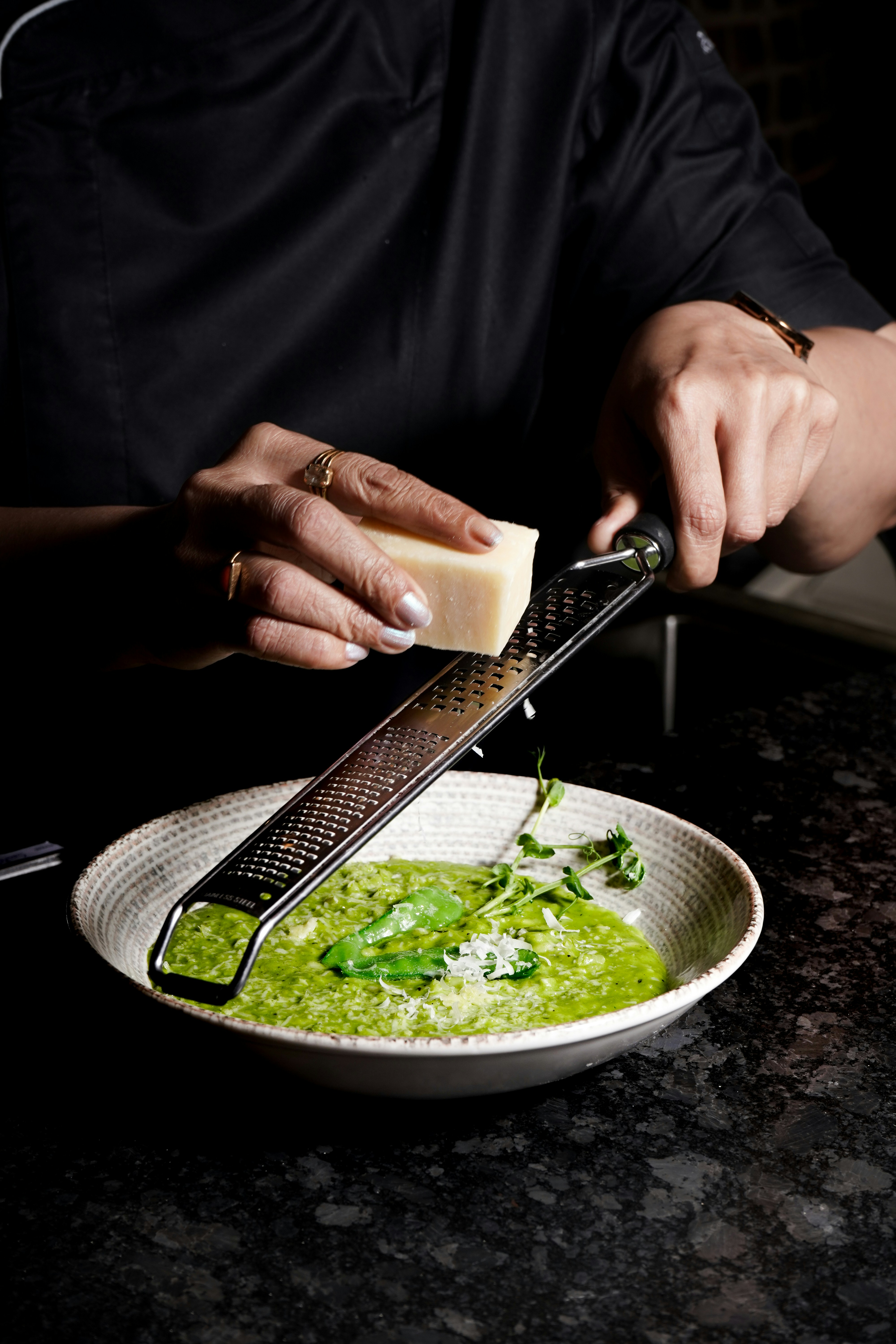 Person reibt Käse über grünes Risotto in weißem Teller